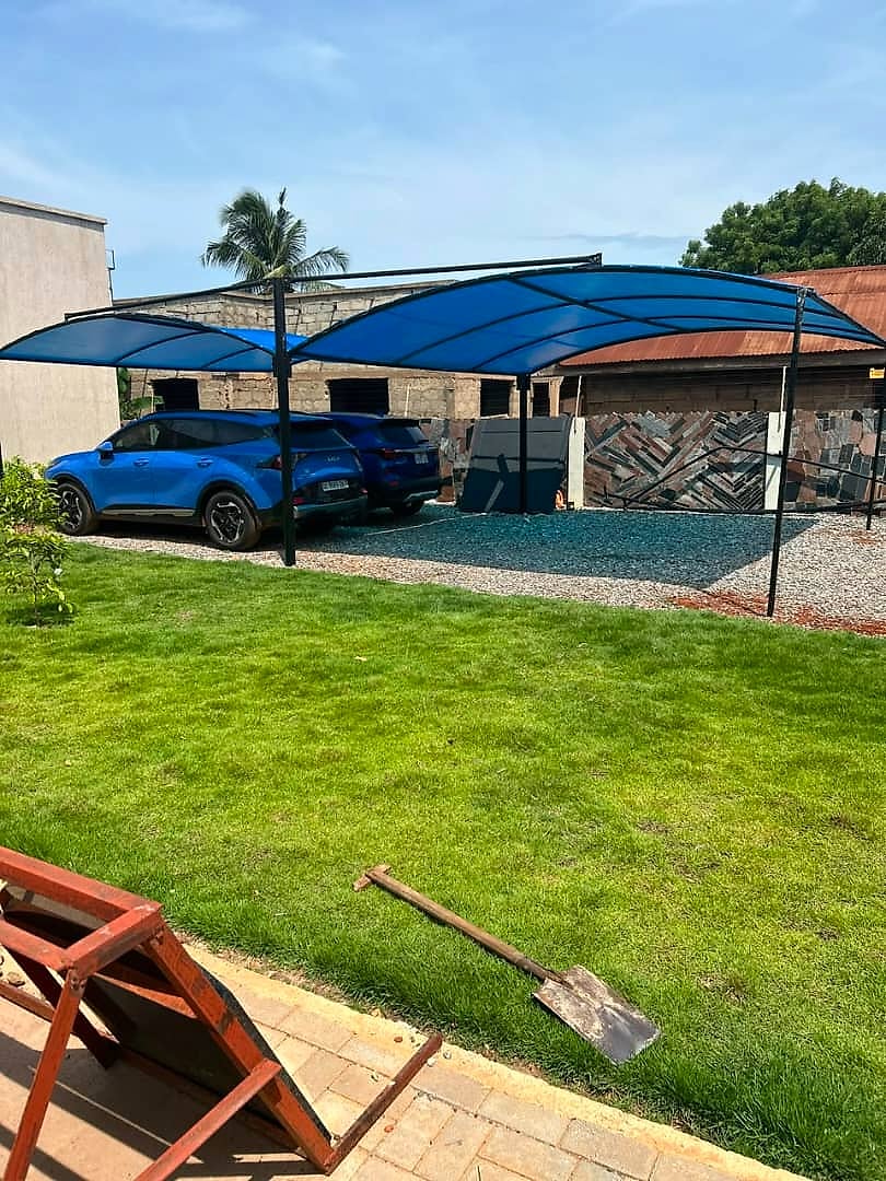 A wide blue polycarbonate carport on black steel posts beside a lawn, with two cars parked underneath