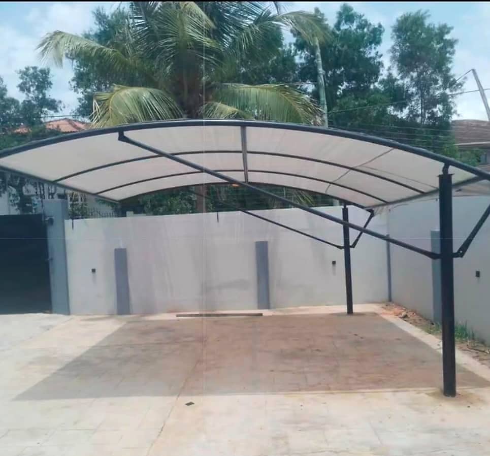 A cream arched shade canopy on a black steel frame inside a walled compound, with a tiled parking area underneath