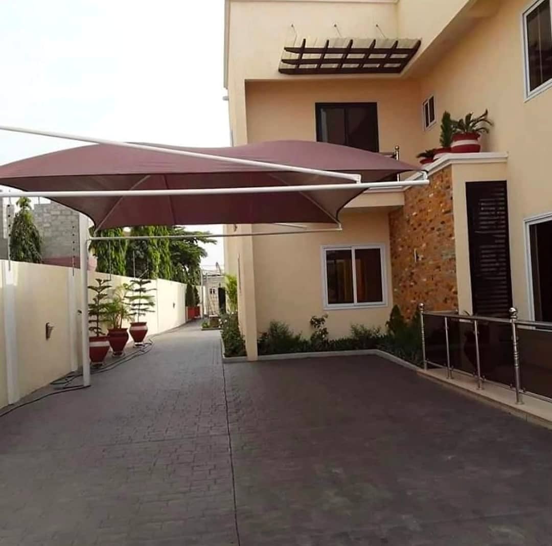 A maroon tensioned-fabric cantilever canopy on white steel arms over a block-paved driveway beside a two-storey house