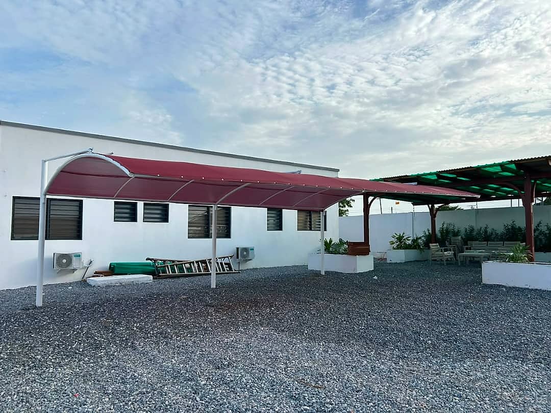 A maroon cantilever shade canopy on white steel posts over a gravelled yard beside a white single-storey building