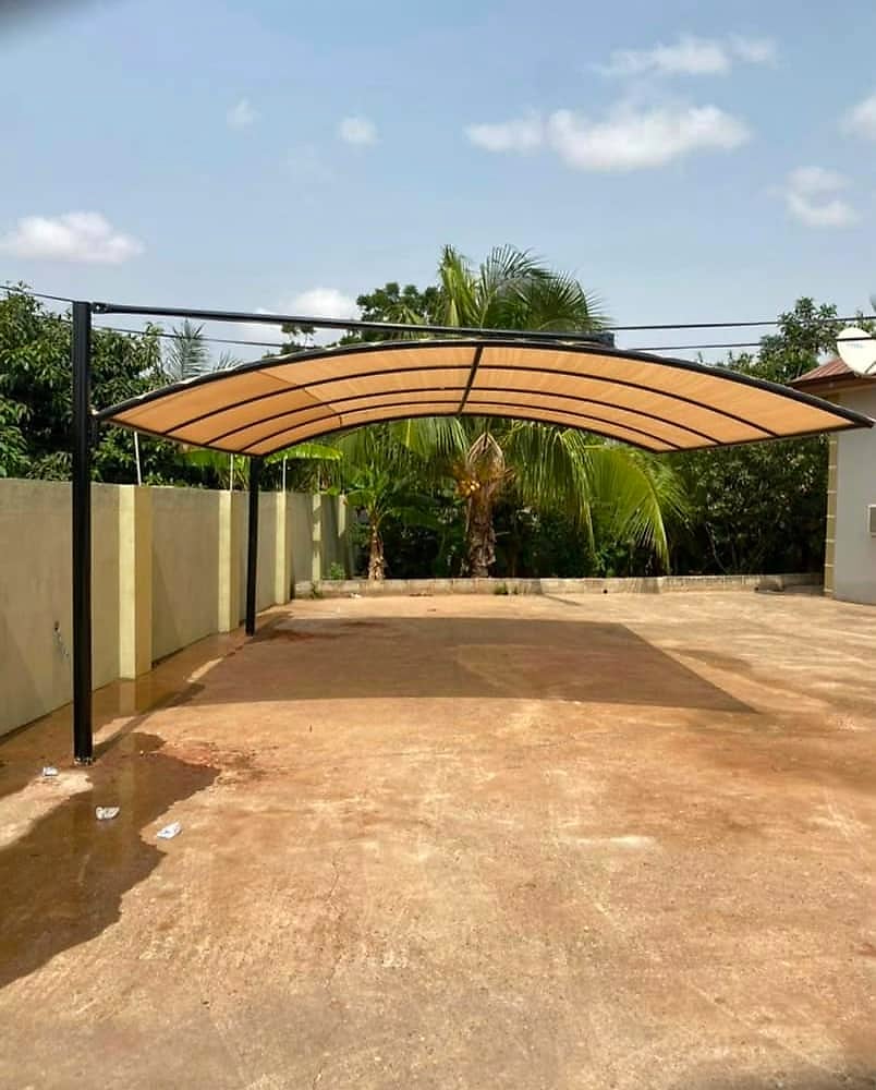 A tan arched shade canopy on a black steel frame standing over an empty compound, with palm trees behind